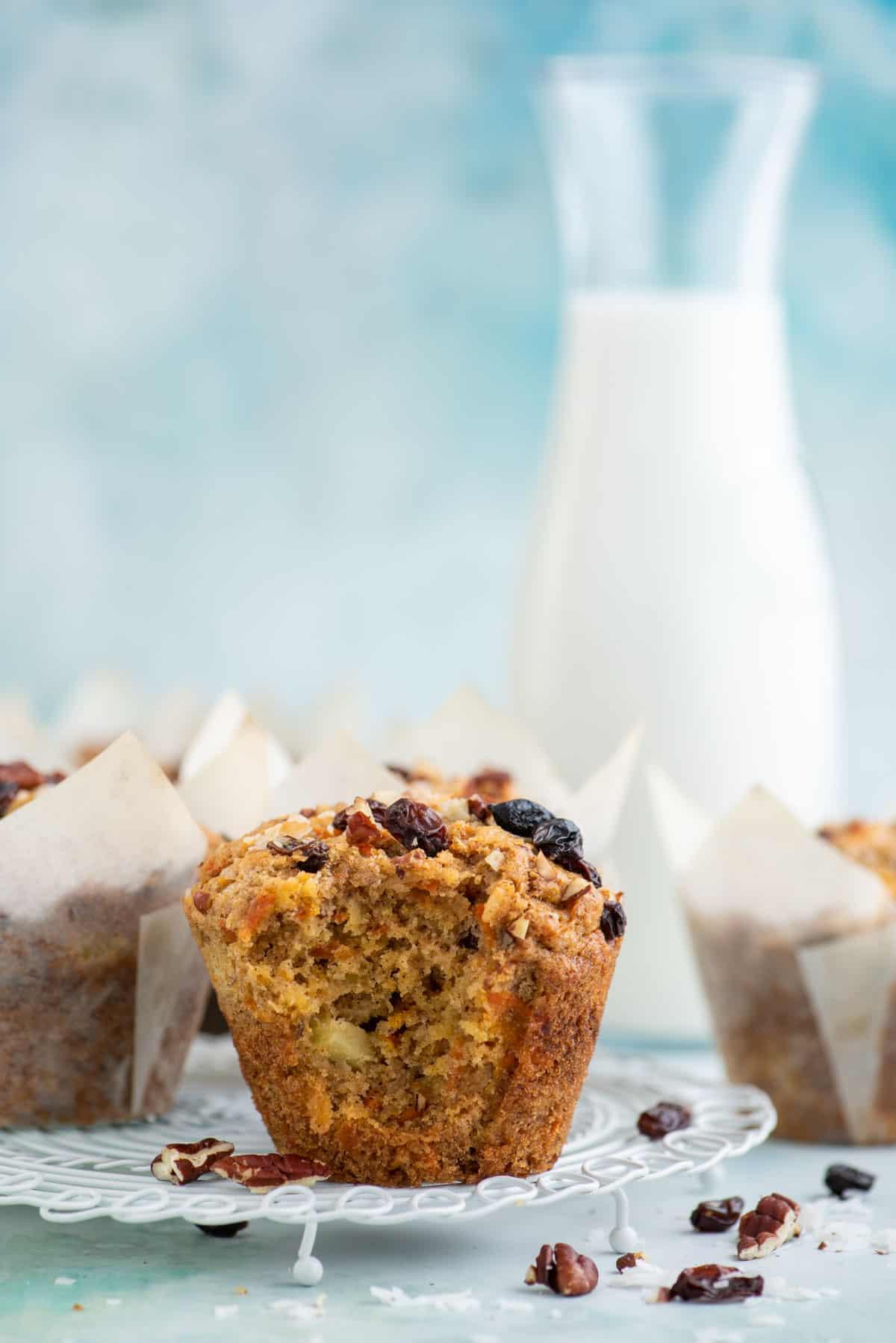 Muffin Morning Glory 4 Un muffin Morning Glory parzialmente mangiato su una griglia bianca, circondato da altri muffin avvolti nella carta, noci, uvetta e scaglie di cocco.