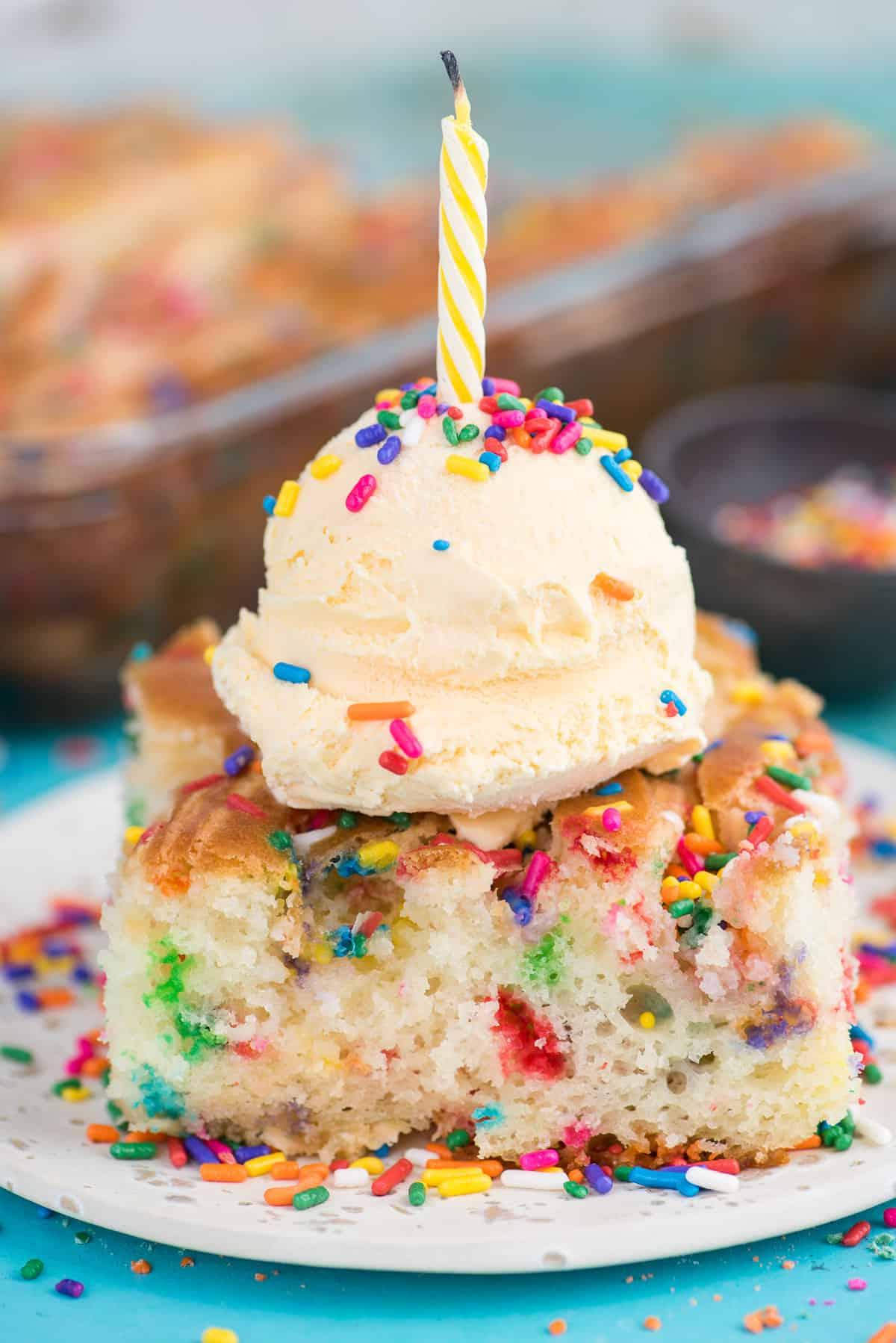 Questa torta terremoto funfetti è un mix per torta bianca arricchito con zuccherini e gocce di cioccolato funfetti. Lo strato di glassa al formaggio cremoso si fonde nella torta creando crepe e fessure in superficie. #earthquakecake #funfetticake #cake Fetta di torta terremoto funfetti con pallina di gelato alla vaniglia su piatto bianco su sfondo blu