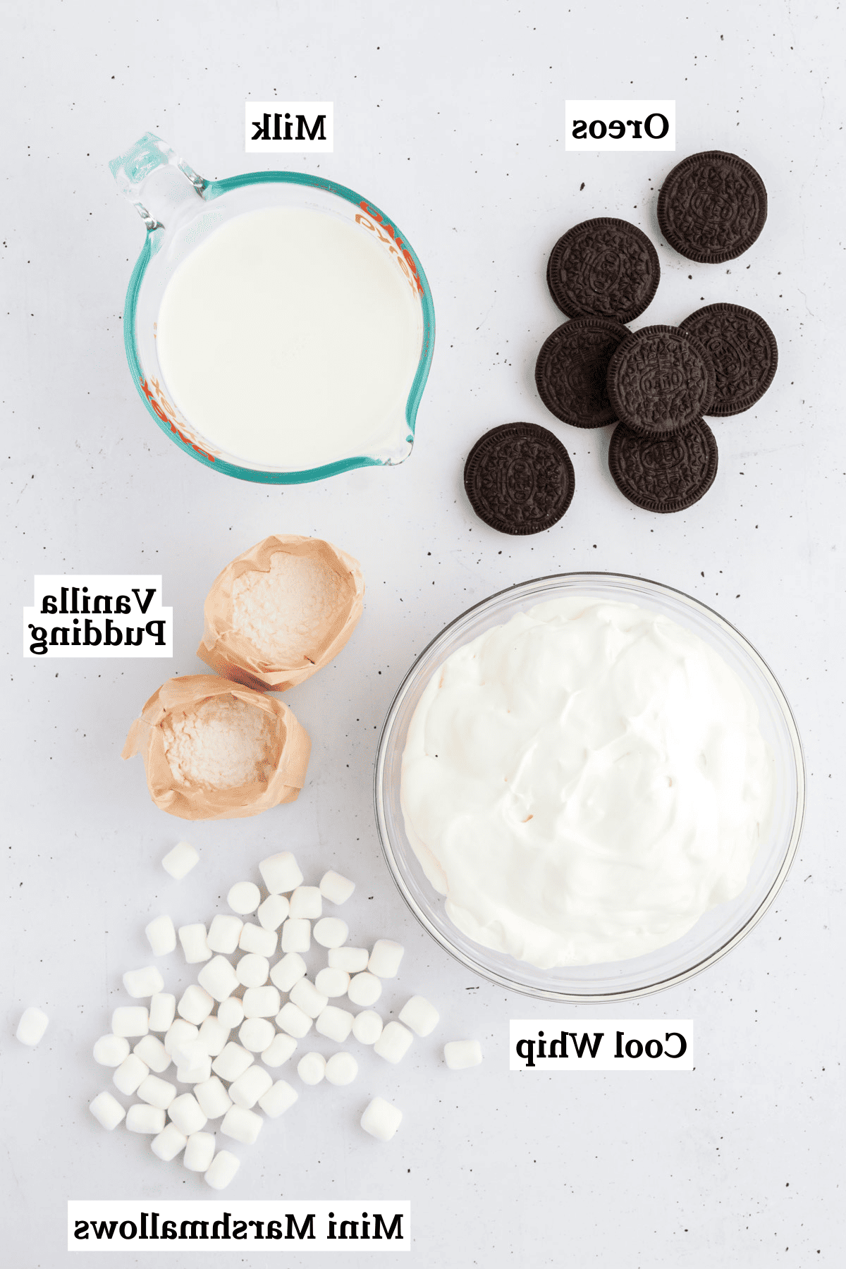 Ricetta Crema Soffice Oreo (Senza Formaggio Cremoso) 4 Ingredienti per il fluff di Oreo disposti su una superficie bianca, inclusi preparato per budino alla vaniglia, latte, Cool Whip, Oreo e mini marshmallow