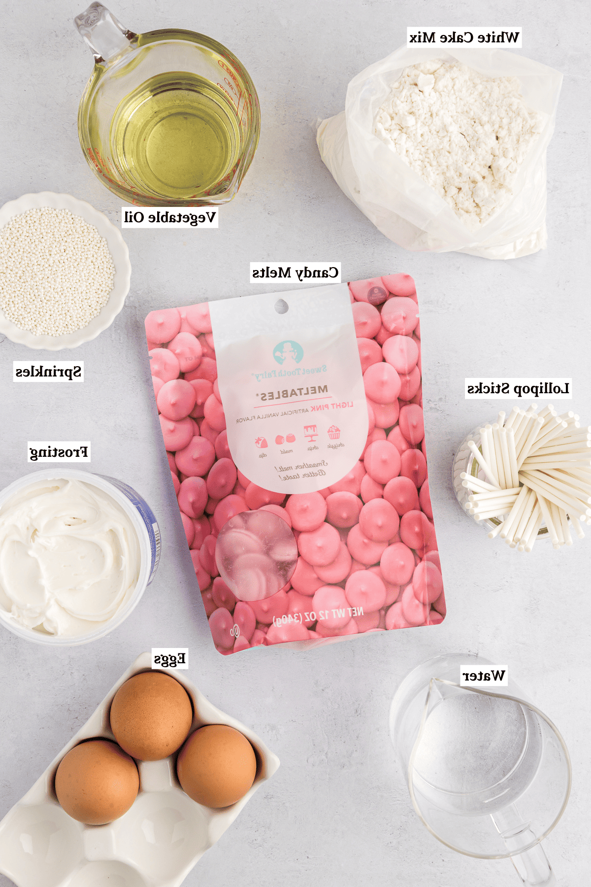 vista dall'alto degli ingredienti per cake pops che includono una busta di preparato per torta bianca, una brocca di misurazione in vetro con olio vegetale, bastoncini per lecca-lecca, una busta di cioccolato fuso rosa, una ciotola bianca di codette bianche, una lattina di glassa bianca, una tazza trasparente di acqua e tre uova