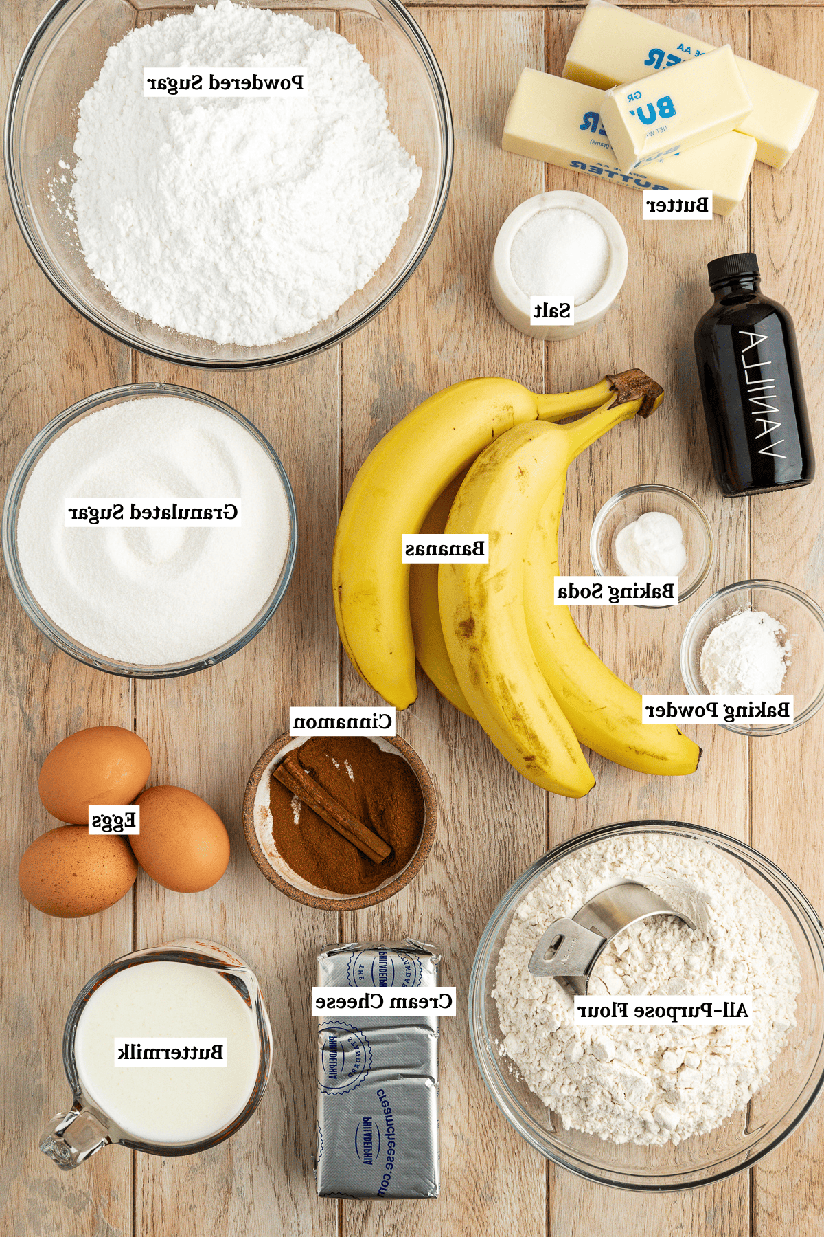 Ricetta Facile Torta alla Banana 4 Vista dall'alto degli ingredienti per la torta di banane su una superficie di legno, tra cui una ciotola di zucchero a velo, una ciotola di zucchero semolato, due bastoncini e mezzo di burro, una piccola ciotola bianca di sale, una bottiglia di estratto di vaniglia, una piccola ciotola di bicarbonato di sodio, una piccola ciotola di lievito in polvere, una ciotola di cannella, tre uova grandi, quattro banane intere, una ciotola di farina per tutti gli usi, un blocco di formaggio cremoso e una tazza misurino di latticello