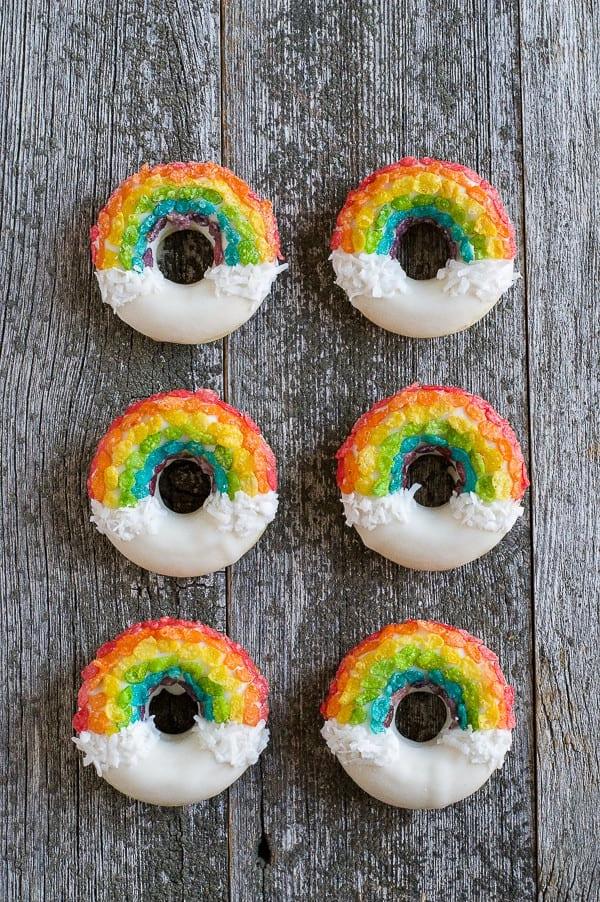Ciambelle Arcobaleno - queste divertenti ciambelle sono fatte con cereali colorati, cioccolato bianco e cocco grattugiato! Non potrai fare a meno di queste deliziose ciambelle! Sei Ciambelle Arcobaleno fatte in casa disposte su un tavolo di legno.