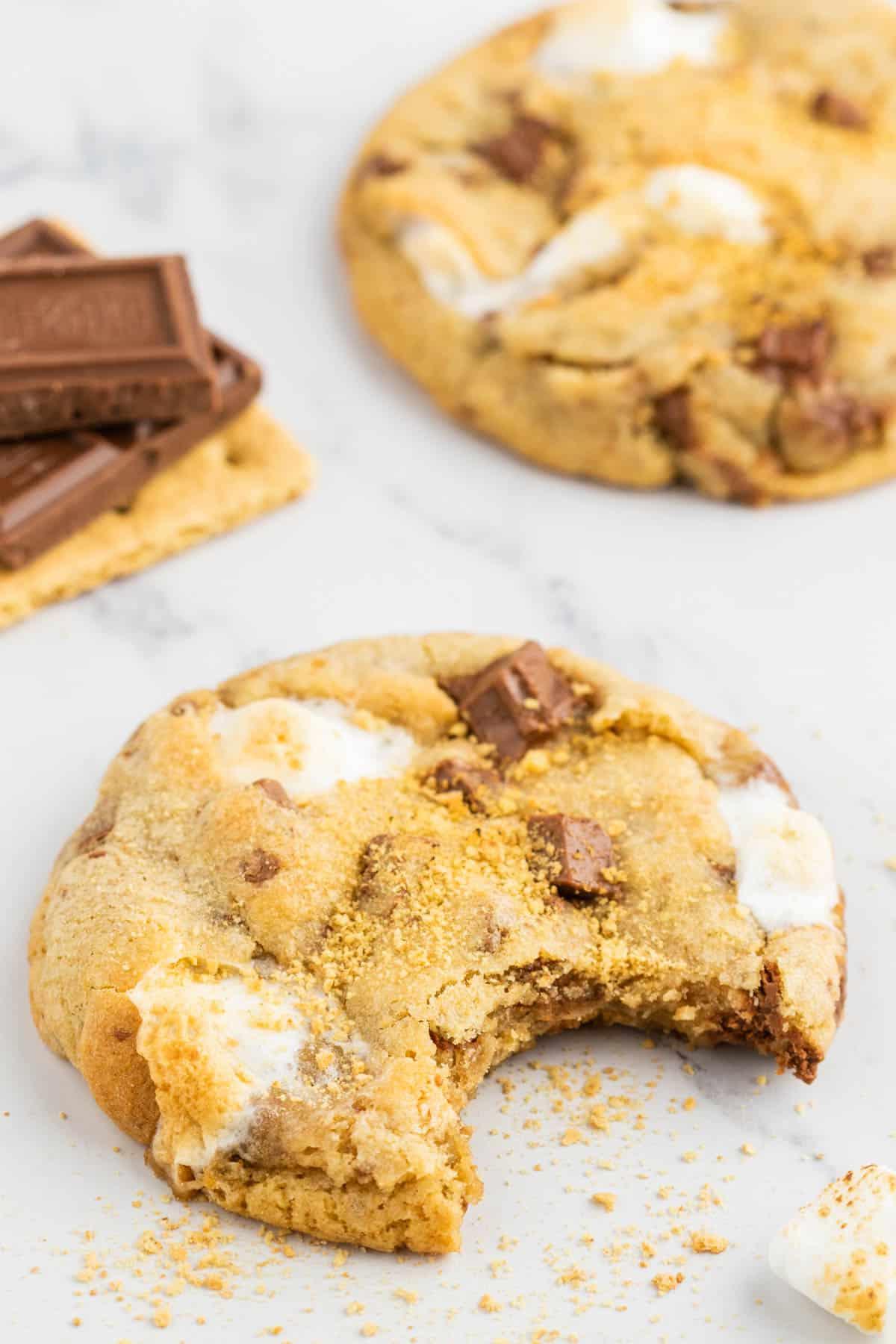 Ricetta Biscotti S'mores con Graham Crackers 4 un biscotto s'mores con un morso e briciole intorno, un altro biscotto sullo sfondo e una pila di cracker Graham con cioccolato Hershey sopra