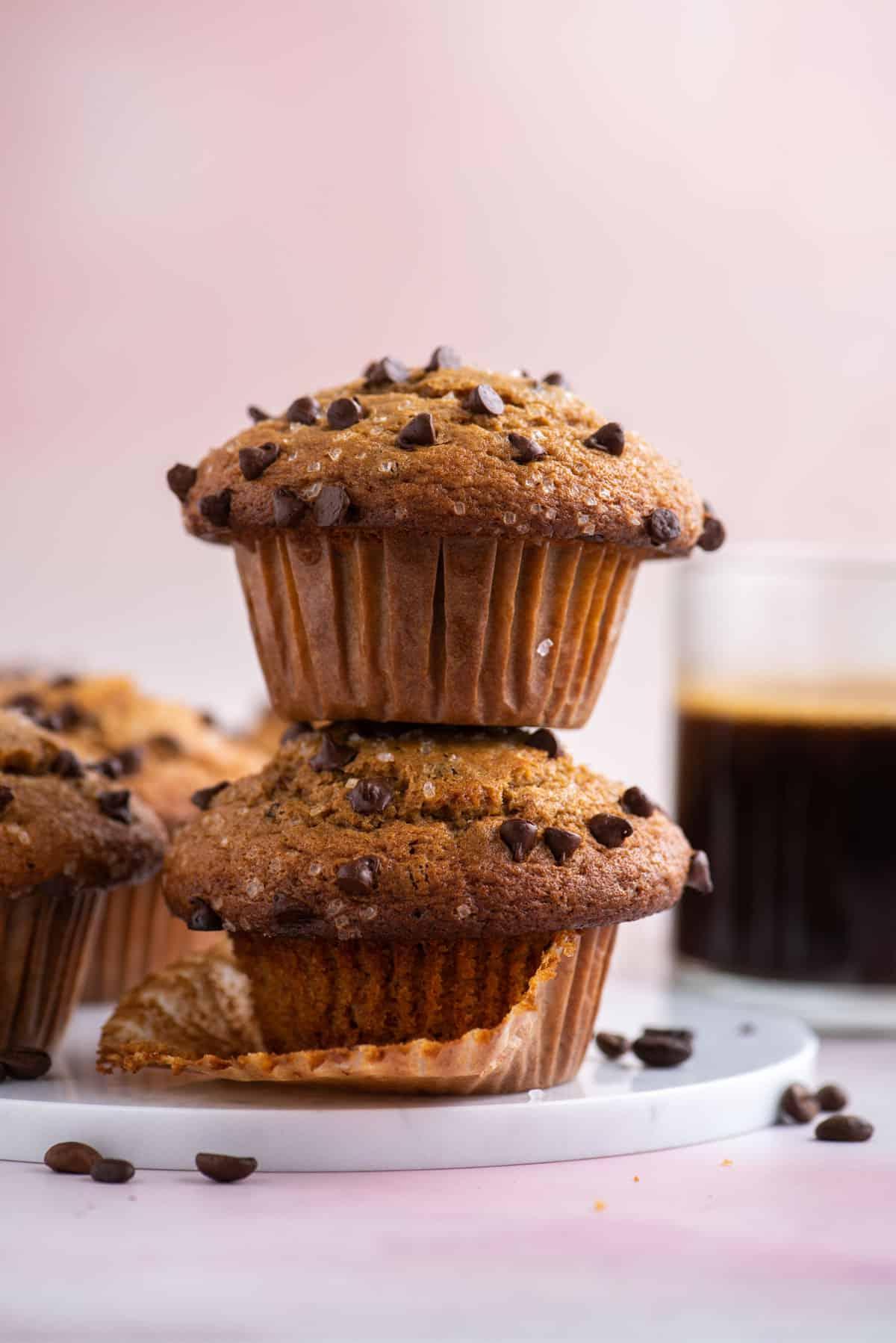 una pila di due muffin al cappuccino con gocce di cioccolato con chicchi di caffè sparsi intorno, altri muffin e una tazza di caffè sullo sfondo