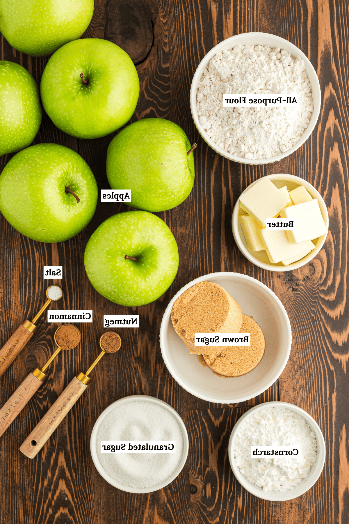 Ricetta Facile di Crumble alle Mele 4 Ingredienti per il crumble di mele disposti su una superficie di legno, tra cui mele verdi, farina 00, burro, zucchero di canna, sale, cannella, noce moscata, zucchero semolato e amido di mais