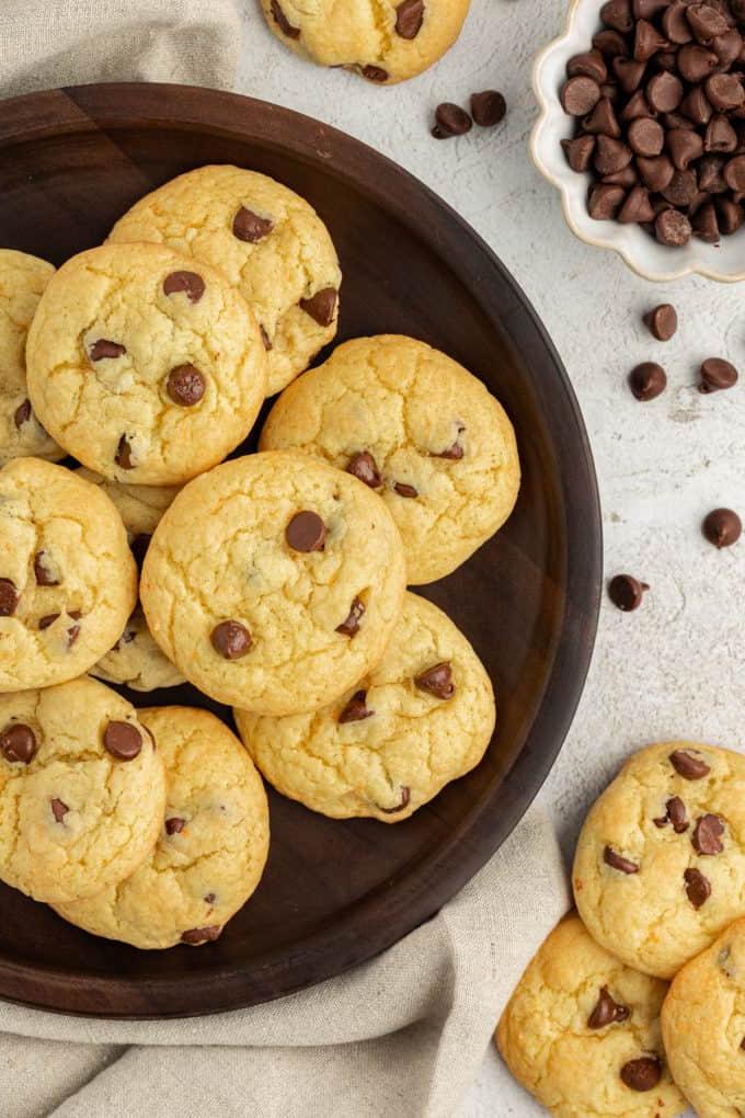 Fare Biscotti dal Mix per Torte 31 Biscotti con gocce di cioccolato e preparato per torte su un piatto marrone su un canovaccio di lino beige, circondati da gocce di cioccolato sparse, altri biscotti e una piccola ciotola bianca di gocce di cioccolato