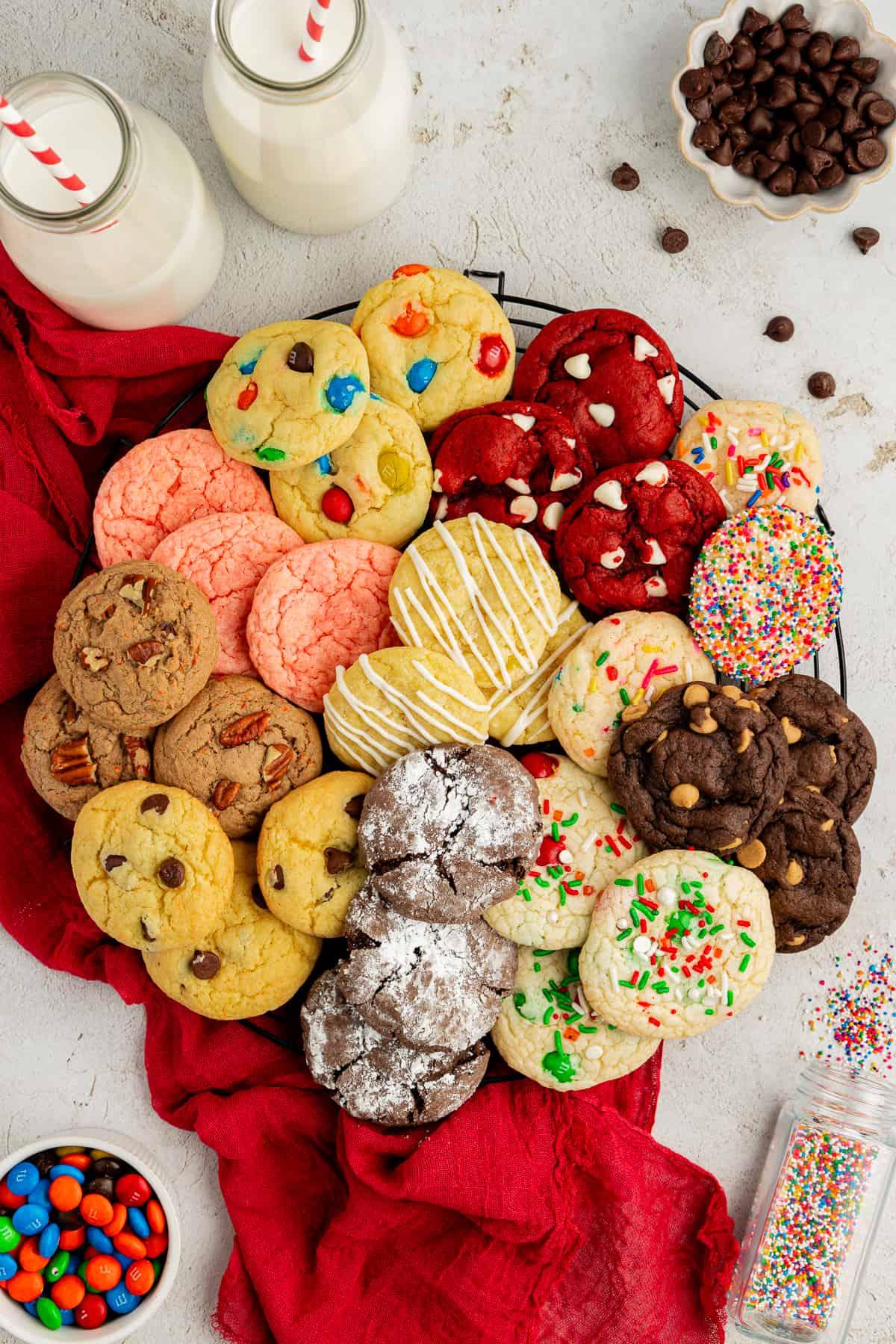 Fare Biscotti dal Mix per Torte 38 Un assortimento di biscotti con preparato per torte di diversi gusti in cerchio su una griglia su un canovaccio rosso con codette, M&M's, gocce di cioccolato e due bicchieri di latte con cannucce rosse e bianche intorno