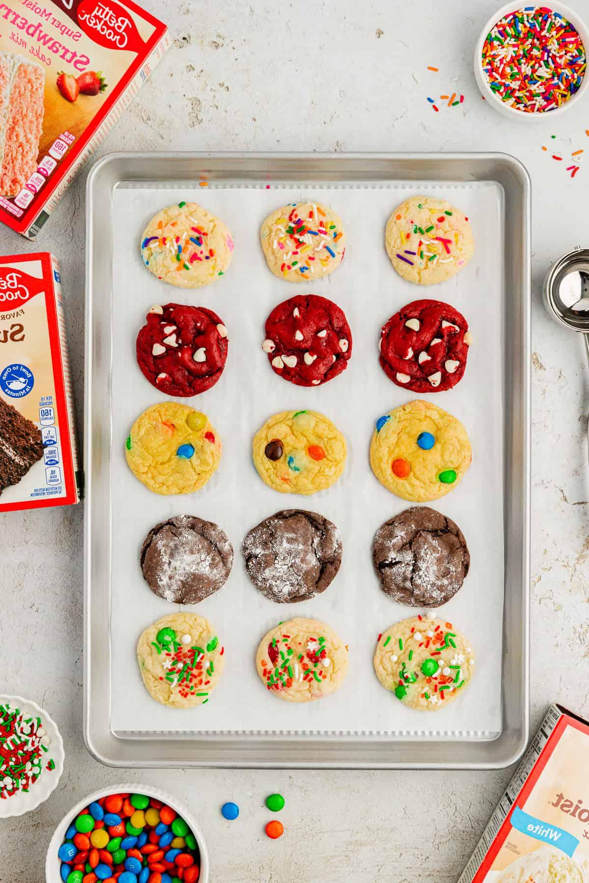 Fare Biscotti dal Mix per Torte 40 Assortimento di biscotti con preparato per torte di diversi gusti in file su una teglia rivestita di carta forno circondata da scatole di preparato per torte, codette colorate, M&M's e uno scoop per biscotti