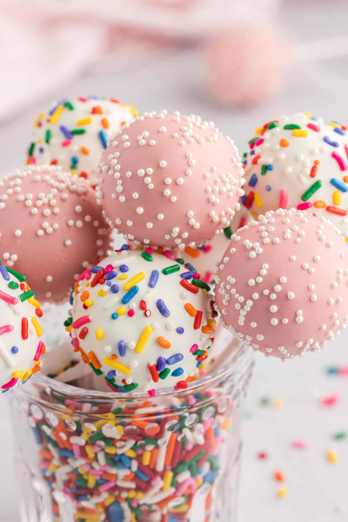 un barattolo di vetro trasparente pieno di codette arcobaleno con cake pops infilati in cima, alcuni bianchi con codette arcobaleno e alcuni rosa con codette bianche