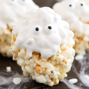 Palle di Popcorn Fantasma 5 Una palla di popcorn fantasma ricoperta di glassa bianca e decorata con due occhietti di zucchero, che assomiglia a una spettrale palla di popcorn di Halloween. Il dolcetto è posato su una superficie nera con sottili filamenti bianchi che ricordano ragnatele.
