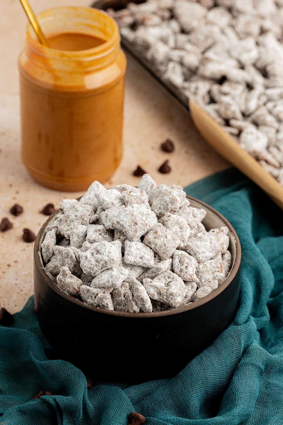 Una ciotola nera piena di puppy chow su un asciugamano color ottanio scuro con un barattolo di burro d'arachidi accanto e una teglia piena di altro puppy chow, con gocce di cioccolato sparse intorno