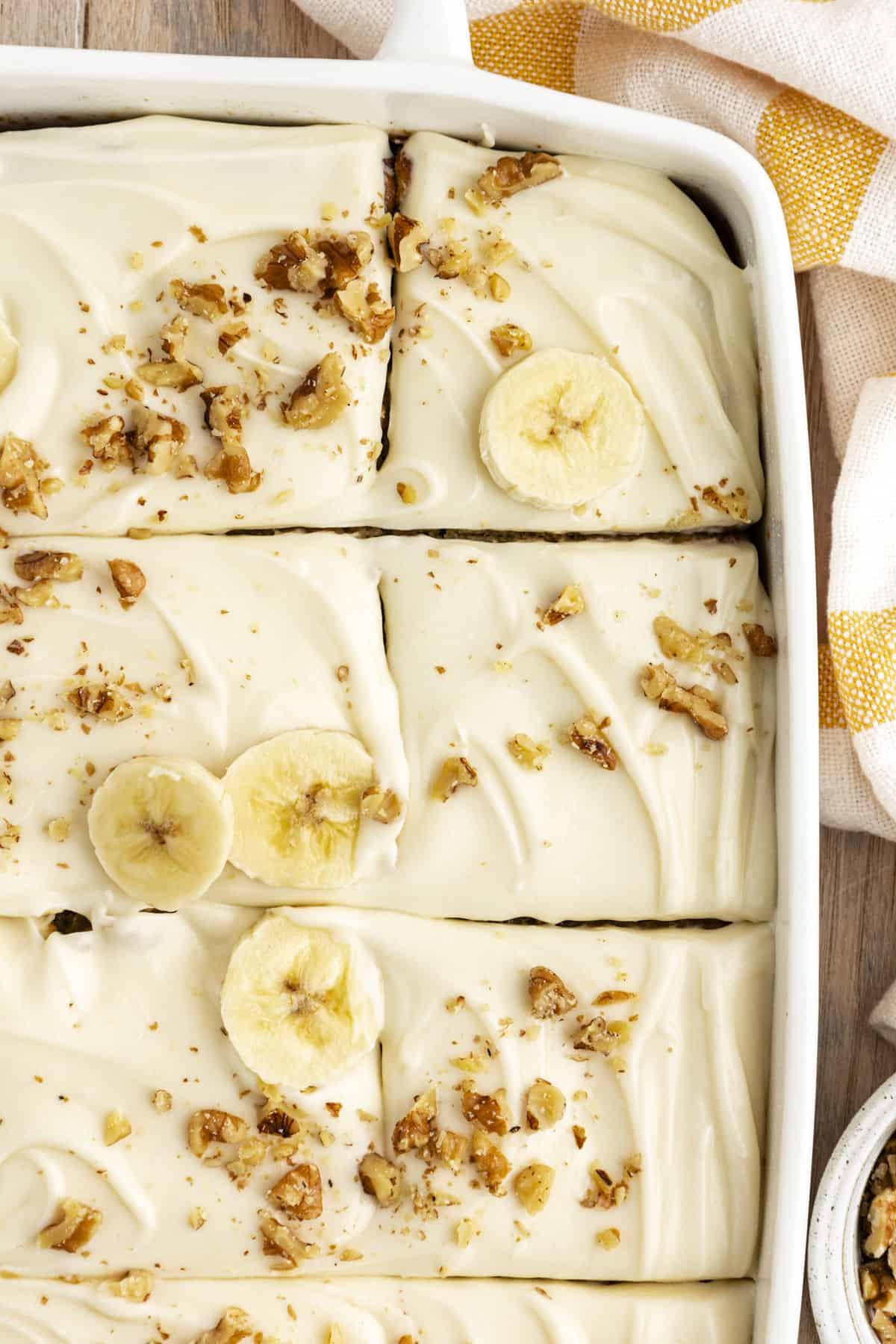 Ricetta Facile Torta alla Banana 5 Vista dall'alto di una torta di banane in una teglia bianca, tagliata a pezzi e guarnita con fette di banana e noci tritate, con un canovaccio da cucina a strisce gialle e bianche accanto alla teglia