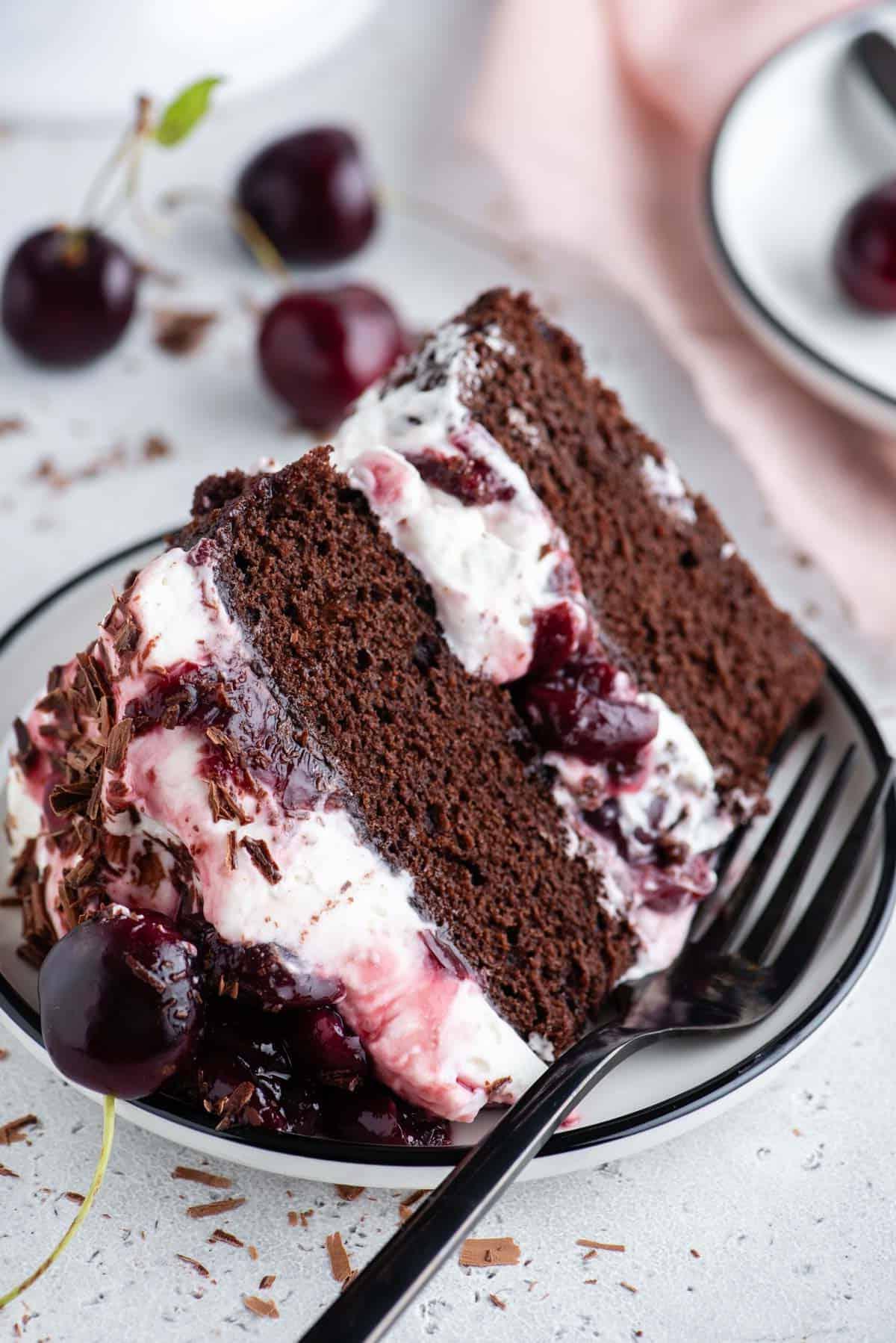 Torta Foresta Nera 5 Una fetta di torta Foresta Nera su un piatto con una forchetta, circondata da ciliegie e piccoli pezzi di cioccolato a scaglie