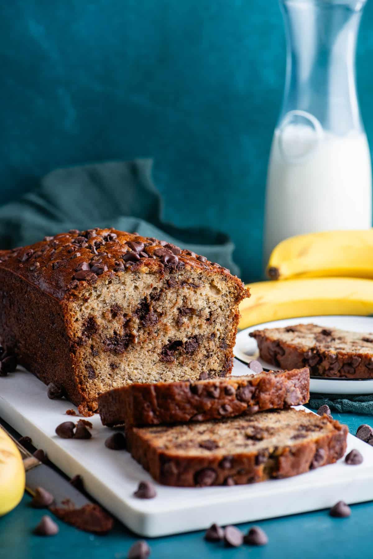 Il Miglior Pan di Banane con Gocce di Cioccolato 5 Una pagnotta di pane alle banane e gocce di cioccolato su un tagliere bianco con un piatto con una fetta di pane alle banane accanto, gocce di cioccolato sparse intorno, e banane intere e un bicchiere di latte sullo sfondo.