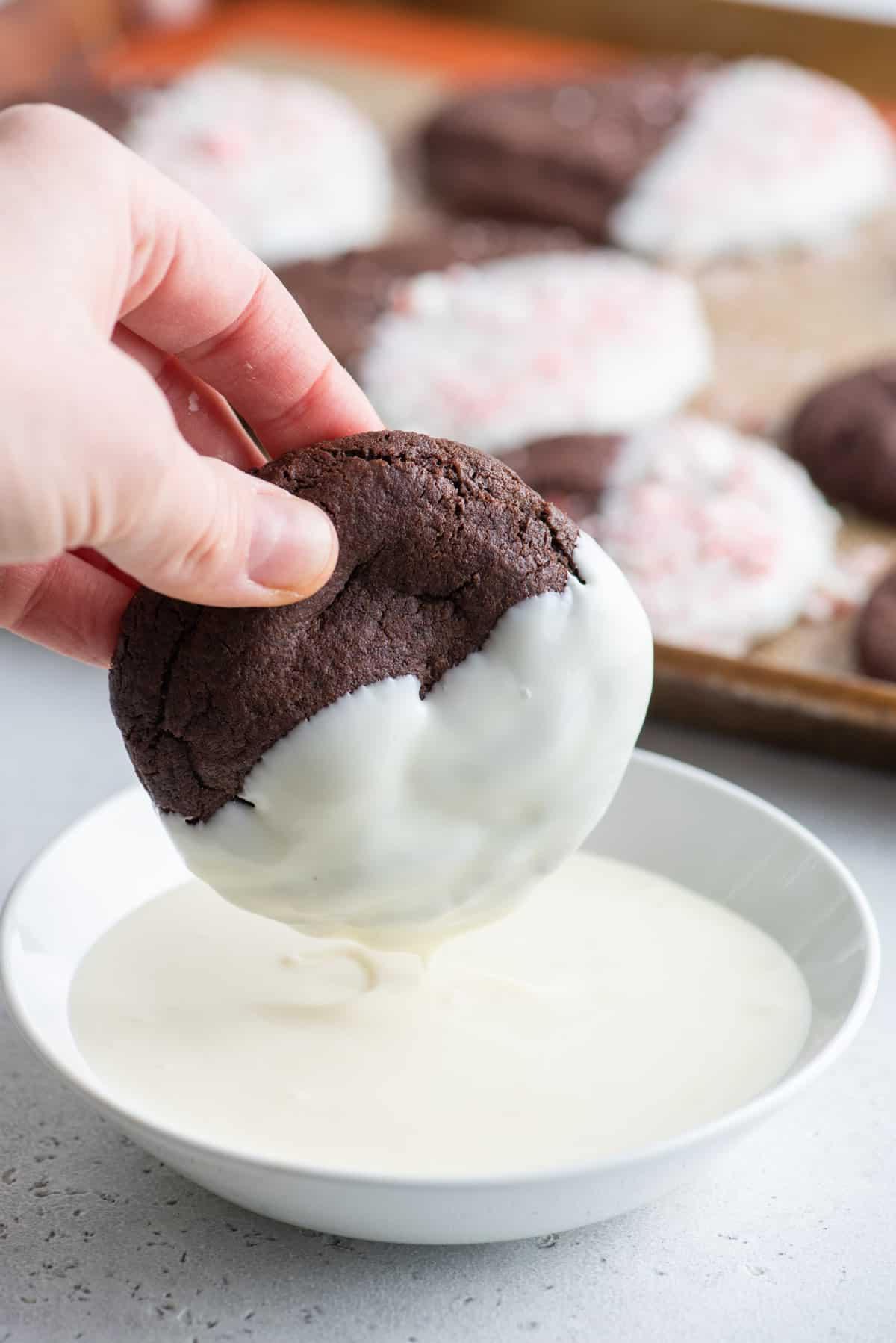 Questi Biscotti al Cioccolato e Bastoncini di Zucchero sono morbidi, gommosi, immersi nel cioccolato bianco e cosparsi di bastoncini di zucchero tritati. Una mano che intinge un biscotto al cioccolato nel cioccolato bianco fuso