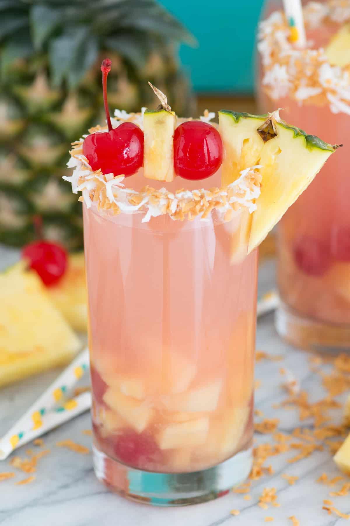 Sangria Piña Colada super facile e tropicale con 5 ingredienti è una bevanda estiva rinfrescante! Questo punch alcolico di sangria ne fa abbastanza per servire una folla. #pinacoladasangria #sangria Sangria Piña Colada in un bicchiere da cocktail con ananas e ciliegia