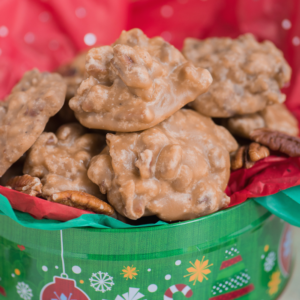 Praline al Pecan 5 Una festosa scatola verde piena di ricche praline di pecan poggia su carta velina rossa e verde. Le praline di pecan dorate e grosse sono lucide e circondate da noci pecan intere, con decorazioni natalizie stampate sulla scatola.