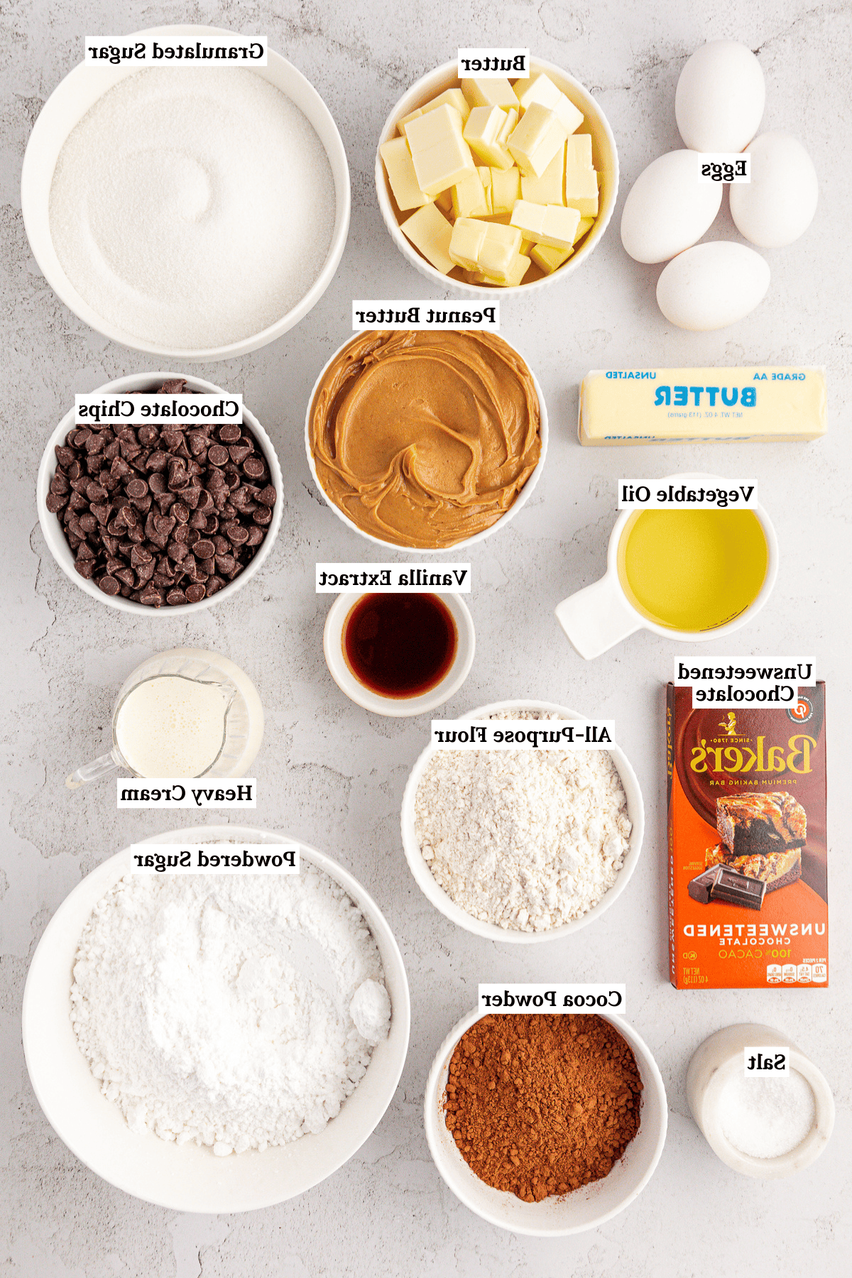 Una varietà di ingredienti per la cottura sono disposti in ciotole e sul bancone, inclusi uova, burro, zucchero semolato, burro d'arachidi, gocce di cioccolato, olio, farina, panna fresca, cioccolato non zuccherato, cacao in polvere, zucchero a velo, vaniglia e sale.