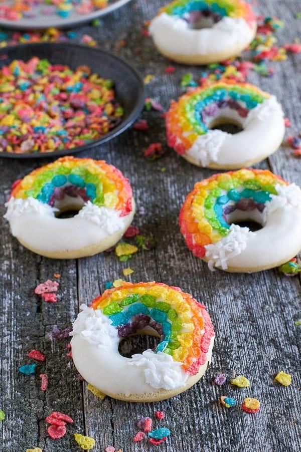 Ciambelle Arcobaleno - queste divertenti ciambelle sono fatte con cereali colorati, cioccolato bianco e cocco grattugiato! Non potrai fare a meno di queste deliziose ciambelle! Quattro semplici Ciambelle Arcobaleno circondate da cereali Fruity Pebbles colorati su un tavolo di legno.