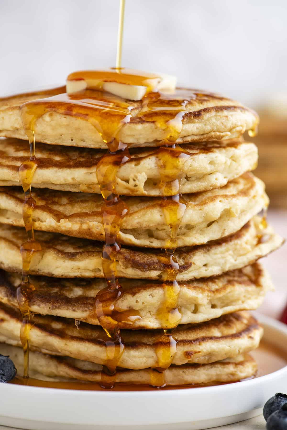 Pancakes Soffici Fatti in Casa 5 Una torre alta di pancake su un piatto bianco, condita con sciroppo, sormontata da una fetta di burro e circondata da frutti di bosco freschi.