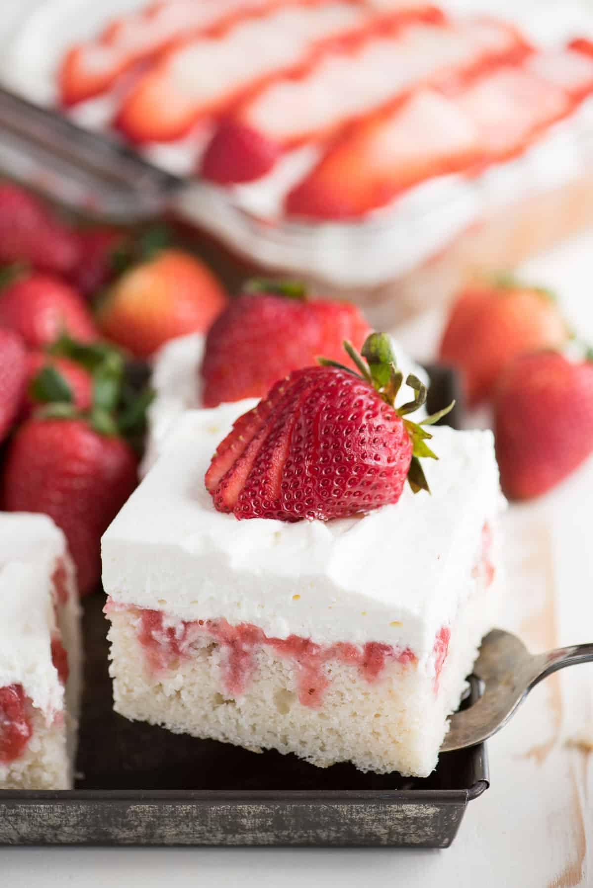 Poke Cake alle Fragole Fresche 5 Una torta poke alle fragole splendidamente decorata, con fragole fresche intere e tagliate, adagiate su una nuvola di panna montata.