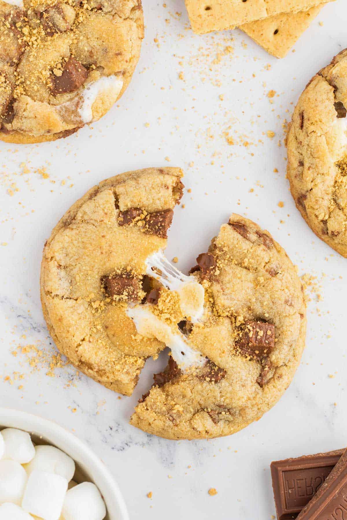 Ricetta Biscotti S'mores con Graham Crackers 5 un biscotto s'mores diviso a metà che mostra marshmallow e cioccolato filanti, disposti con altri biscotti, cioccolato Hershey, marshmallow e cracker Graham intorno