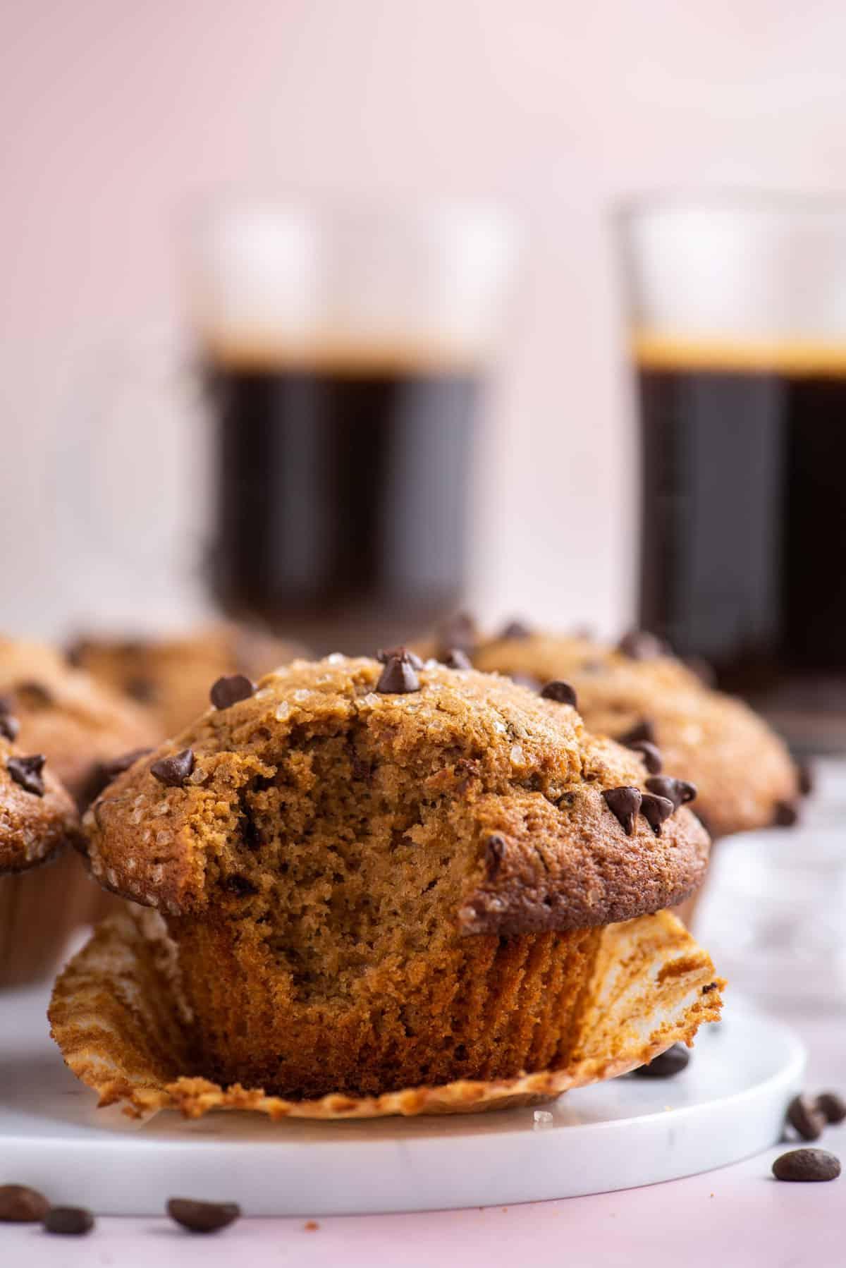 un muffin al cappuccino con gocce di cioccolato su un piatto bianco con un morso preso, appoggiato nel suo pirottino aperto, con chicchi di caffè sparsi intorno, altri muffin e due tazze di caffè sullo sfondo