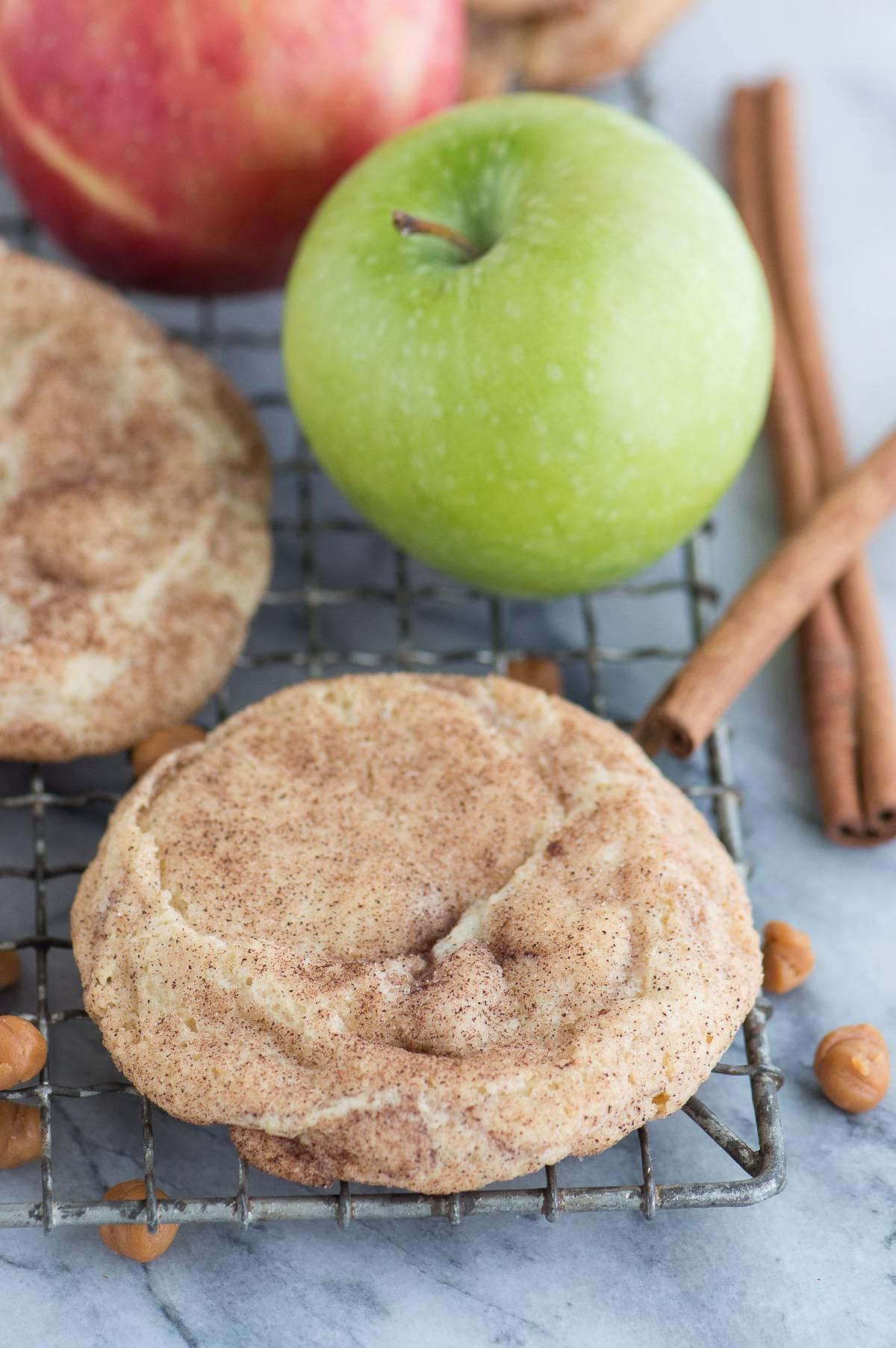 Snickerdoodles al Caramello e Mela 5 Biscotto snickerdoodle alla mela caramellata su una griglia di raffreddamento con caramelle, bastoncini di cannella e una mela sullo sfondo