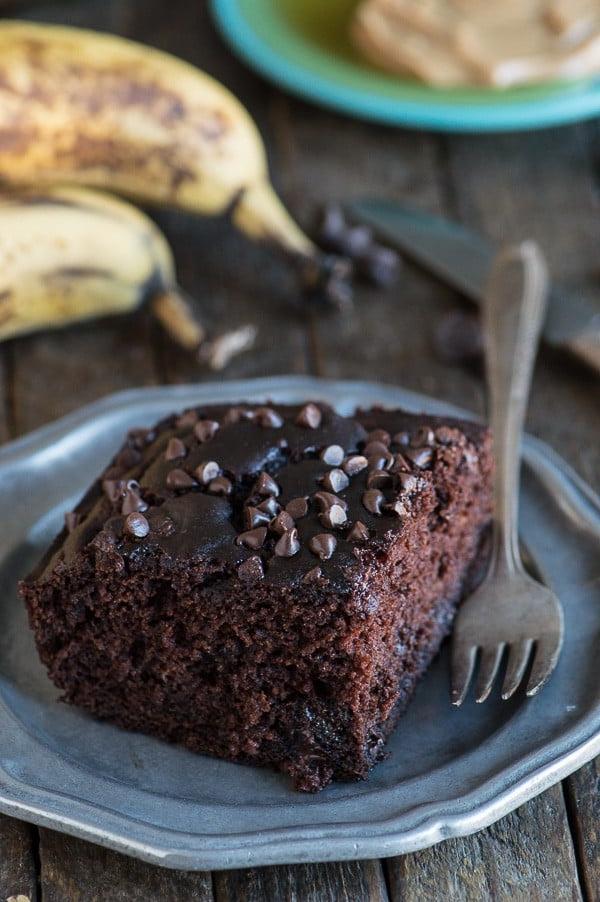 Questa torta al cioccolato più sana sa di muffin alla banana con doppie gocce di cioccolato! Niente zucchero, burro o olio, ma usa banane, yogurt greco e miele invece! Torta al cioccolato salutare su piatto con banane sullo sfondo