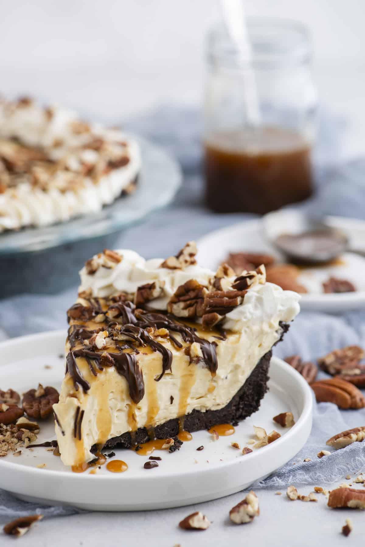 Torta di Tartaruga 5 Una fetta di Torta Tartaruga guarnita con noci pecan, gocce di cioccolato e caramello, su un piccolo piatto bianco con pezzi di noci pecan sparsi intorno.