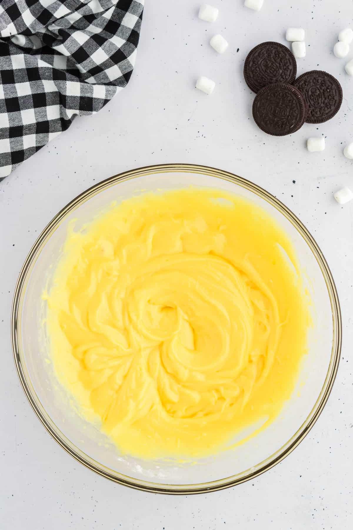 Ricetta Crema Soffice Oreo (Senza Formaggio Cremoso) 5 Budino alla vaniglia in una ciotola di vetro circondato da Oreo, mini marshmallow e un panno da cucina a scacchi bianchi e neri