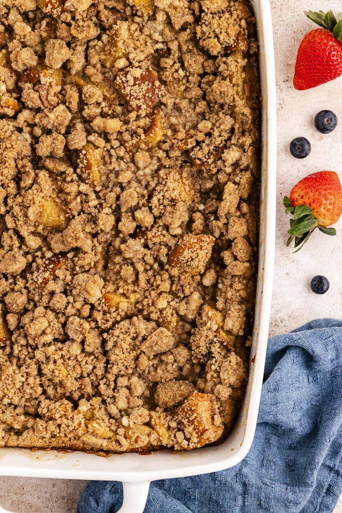Migliore Ricetta per Budino di French Toast 5 Una grande teglia bianca di casseruola di French Toast con topping croccante accanto a un canovaccio blu e mirtilli e fragole fresche sparse intorno.