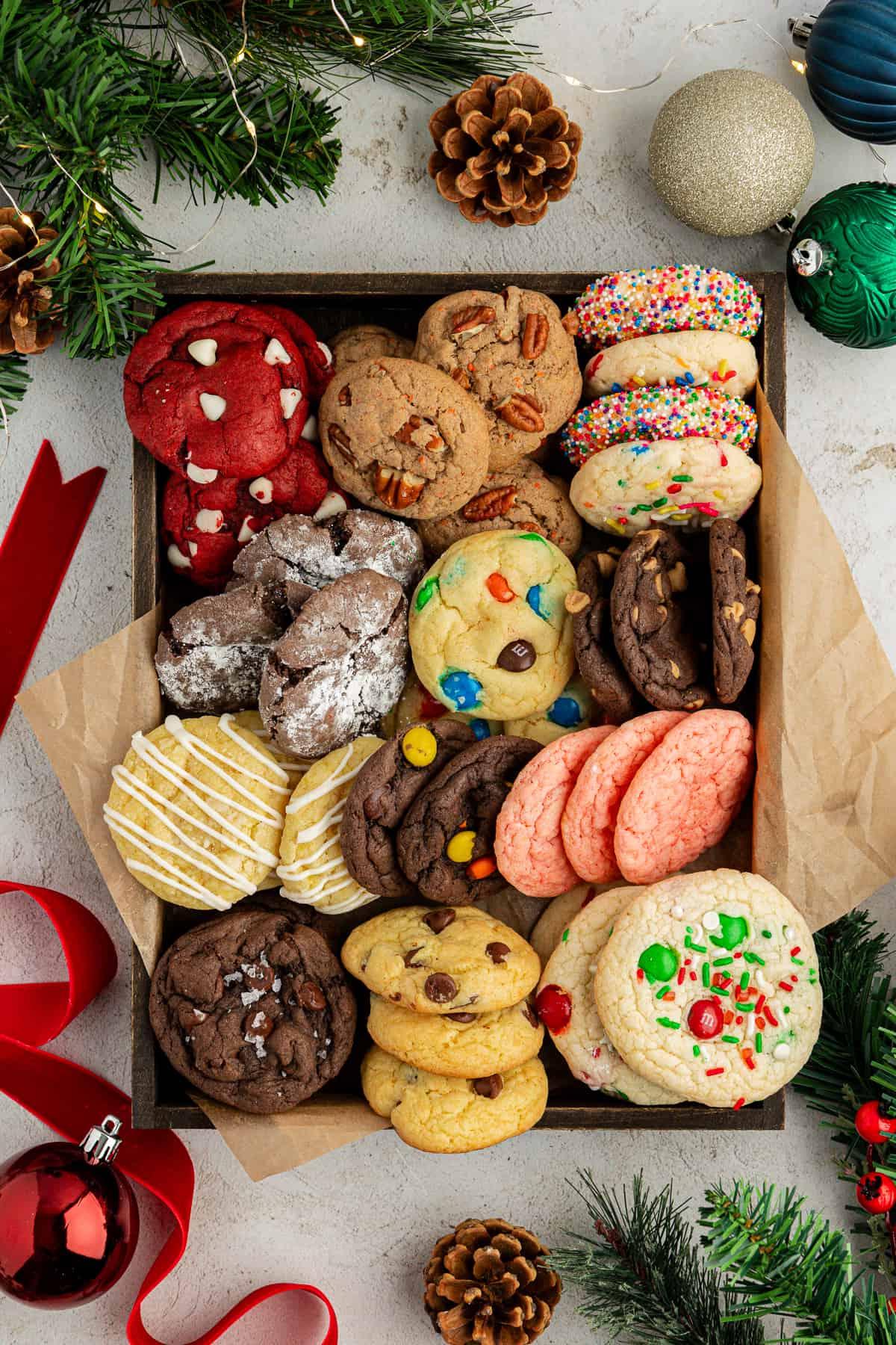 Fare Biscotti dal Mix per Torte 41 Una scatola piena di biscotti con preparato per torte di vari gusti con carta marrone circondata da ornamenti natalizi, nastro rosso, pigne e rami di albero di Natale