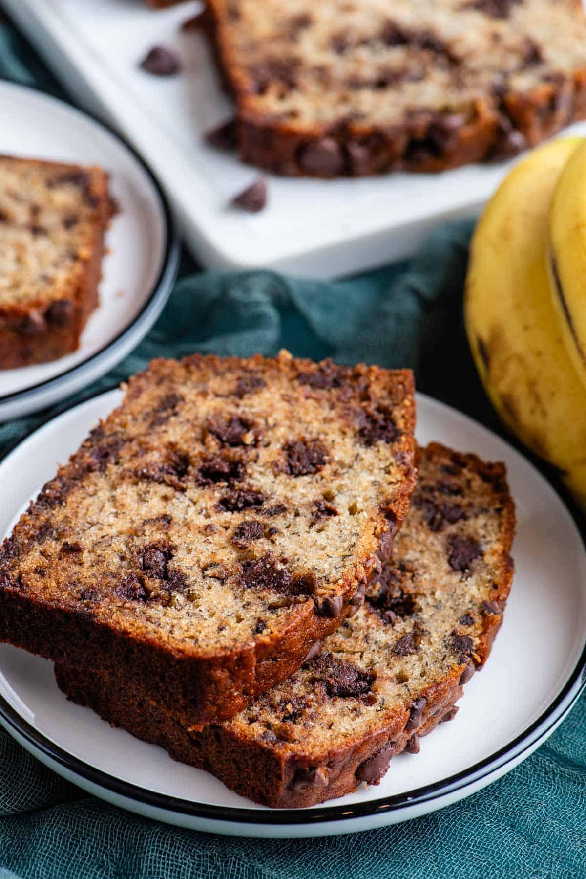 Il Miglior Pan di Banane con Gocce di Cioccolato 6 Due fette di pane alle banane e gocce di cioccolato su un piatto bianco sopra un asciugamano color ottanio scuro, con banane intere accanto, un altro piatto con una fetta di pane alle banane, e un tagliere bianco con altro pane dietro.