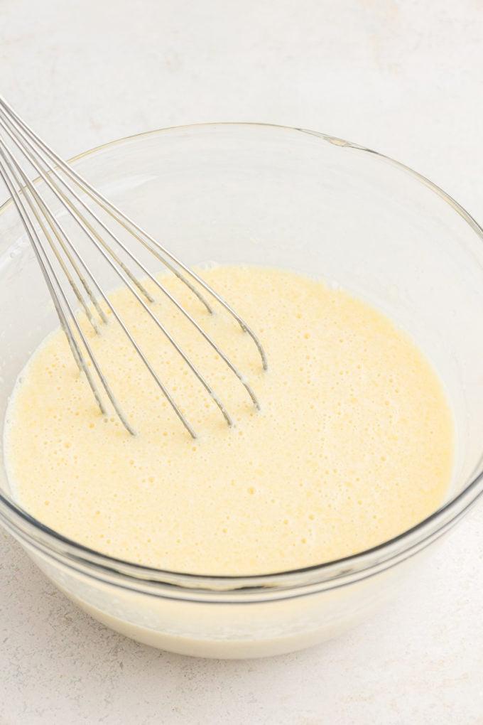 Ricetta Pancakes in Teglia 6 Ingredienti liquidi per pancake al forno in teglia in una ciotola di vetro trasparente con una frusta appoggiata all'interno della ciotola.