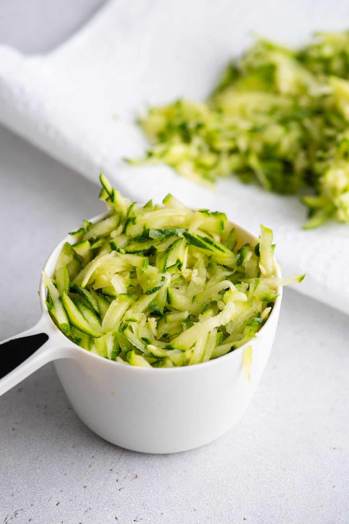 Migliori Muffin alle Zucchine 6 Una tazza di misurazione bianca di zucchine grattugiate accanto a un tovagliolo di carta con più zucchine sopra