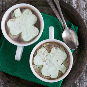 Marshmallow alla Menta e Gocce di Cioccolato al Trifoglio 2 Due tazze di cioccolata calda con marshmallow verdi a forma di trifoglio su un vassoio scuro con due cucchiaini di metallo e un tovagliolo di stoffa verde, il tutto su una superficie di legno rustico.