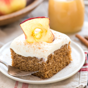 Torta Speziata alla Salsa di Mele 6 Una fetta di torta di mele speziata con glassa bianca, una spolverata di cannella e una fetta di mela essiccata, servita su un piatto bianco con una forchetta. Un bicchiere e della frutta sono sfocati sullo sfondo.
