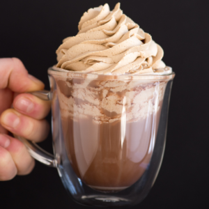 Panna montata al caffè 5 Una mano tiene una tazza di vetro trasparente piena di cioccolata calda, guarnita con un generoso vortice di panna montata al caffè su uno sfondo nero semplice.
