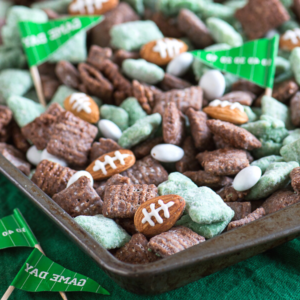 Calcio Puppy Chow 6 Un vassoio pieno di Chex mix verde e marrone, Puppy Chow a tema football, caramelle ovali bianche, snack a forma di pallone da football e piccole bandierine di carta con scritto Game Day, il tutto su una superficie verde scuro.