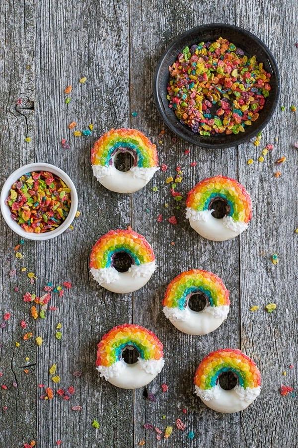 Ciambelle Arcobaleno - queste divertenti ciambelle sono fatte con cereali colorati, cioccolato bianco e cocco grattugiato! Non potrai fare a meno di queste deliziose ciambelle! Sei Ciambelle Arcobaleno accanto a due ciotole di cereali Fruity Pebbles su un tavolo di legno.