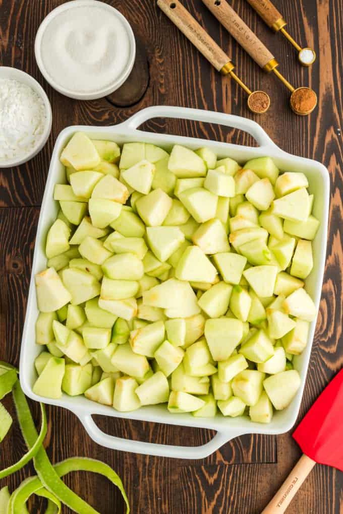 Ricetta Facile di Crumble alle Mele 6 Una pirofila bianca piena di mele verdi tritate su una superficie di legno circondata da misurini e piccole ciotole piene di zucchero, amido di mais, cannella e noce moscata, una spatola rossa e bucce di mela verde