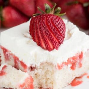 Poke Cake alle Fragole Fresche 6 Una fetta di torta poke alle fragole con salsa di fragole e glassa bianca, guarnita con una fragola a fette, poggia su un piatto bianco con fragole fresche sullo sfondo. Un morso è stato preso dalla torta.