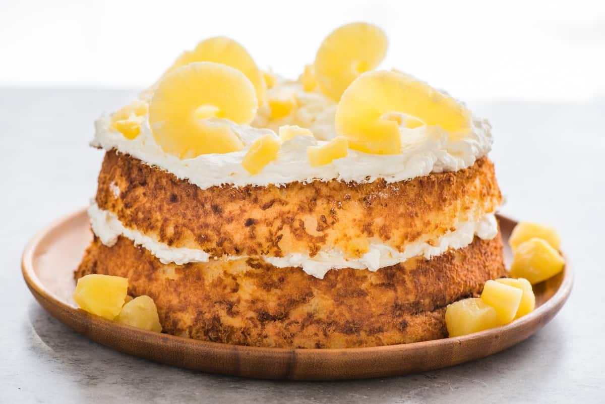 Torta Angel Food intera guarnita con panna montata e fette di ananas su vassoio di legno su sfondo bianco