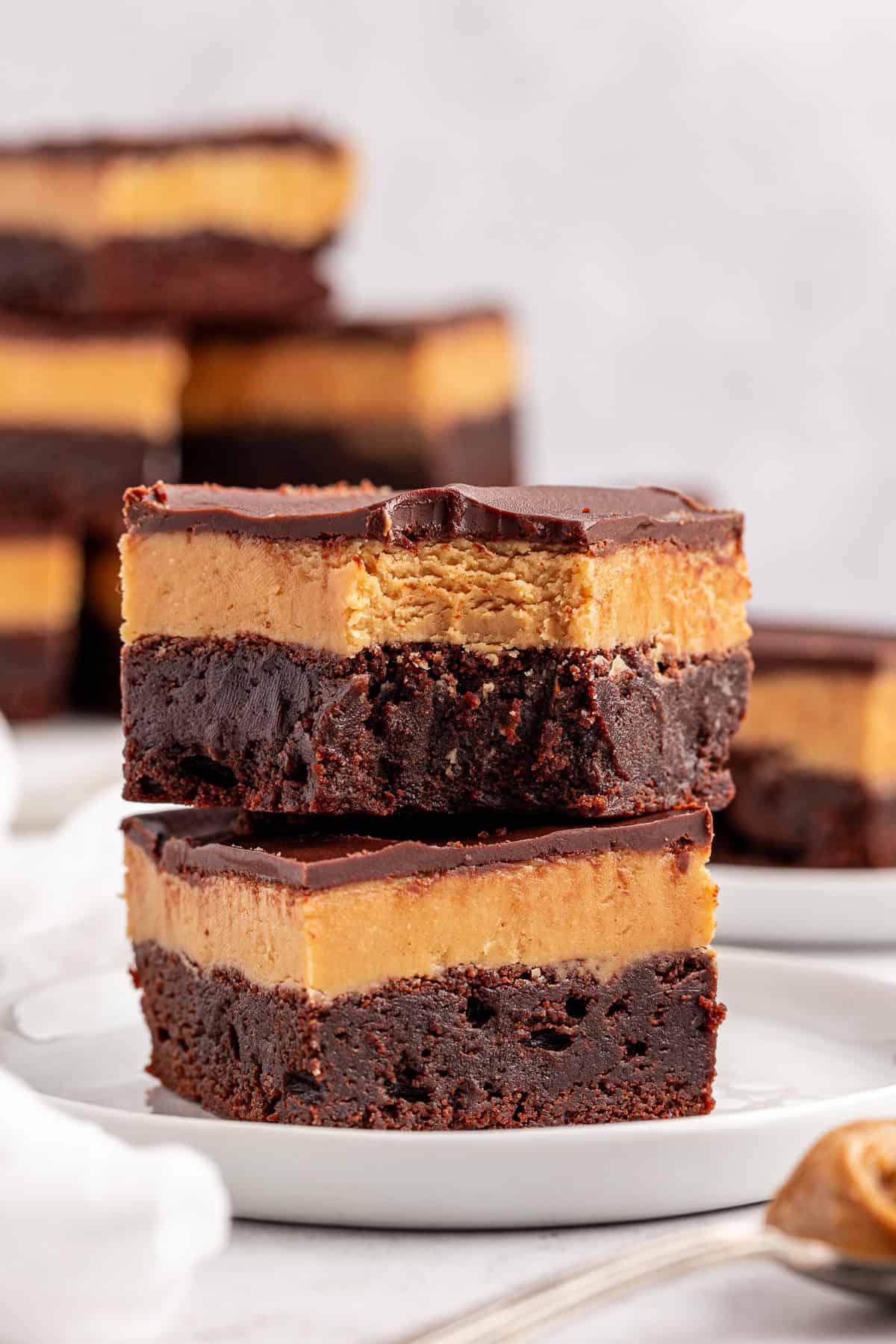 Due brownies Buckeye con uno spesso strato di burro d'arachidi e una copertura di cioccolato sono impilati su un piatto bianco. Altri brownies sono visibili sullo sfondo, leggermente sfocati.