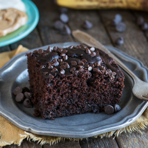 Torta al cioccolato più salutare 3 Una fetta di torta al cioccolato umida e salutare, guarnita con gocce di cioccolato, su un piatto grigio con una forchetta, su un tavolo di legno rustico. Gocce di cioccolato sparse e alimenti sfocati sono visibili sullo sfondo.