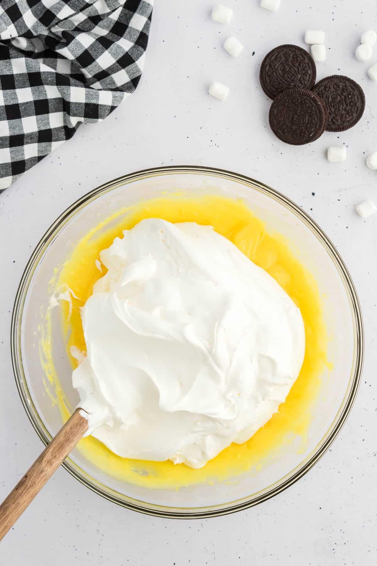 Ricetta Crema Soffice Oreo (Senza Formaggio Cremoso) 6 Budino alla vaniglia in una ciotola di vetro sormontato da Cool Whip, circondato da Oreo, mini marshmallow e un panno da cucina a scacchi bianchi e neri