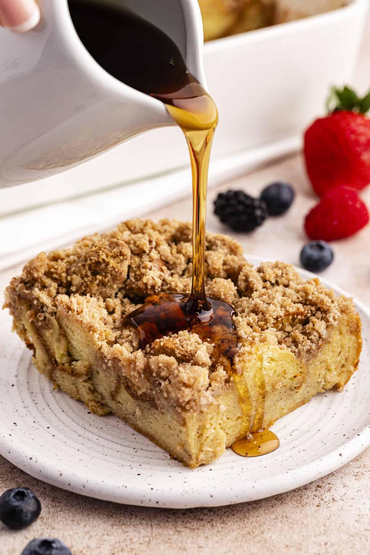 Migliore Ricetta per Budino di French Toast 6 Una fetta di casseruola di French Toast su un piattino con sciroppo d'acero versato sopra e frutti di bosco freschi sparsi intorno.