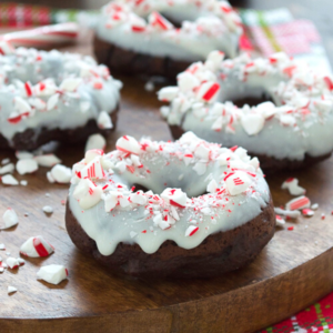 Ciambelle al cioccolato guarnite con glassa bianca e caramelle alla menta piperita tritate, disposte su un tagliere di legno. Alcuni pezzi di ciambella al bastoncino di zucchero e pezzetti di menta sono sparsi intorno, creando un aspetto festivo.