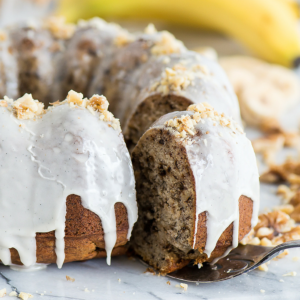 Torta Bundt alle Noci e Banane 7 Una torta Bundt di banana e noci umida con glassa bianca che cola e noci tritate sopra; una fetta viene sollevata, rivelando la morbida consistenza della torta. Banane e altre noci sono sfocate sullo sfondo.